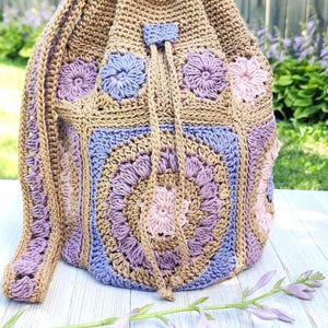 K&ouml;nnte beinhalten: Handgefertigte H&auml;keltasche in Beige-, Lavendel- und Rosat&ouml;nen. Die Tasche hat ein Blumenmuster, einen Kordelzugverschluss und einen Schulterriemen. Der Text "Kathy's Crochet Closet" und "Petals Bucket Bag Crochet Pattern" sind sichtbar.