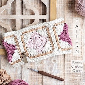May include: A crocheted headband featuring a pink and white flower design with brown accents. The headband is displayed on a wooden surface with a crochet hook and balls of yarn.