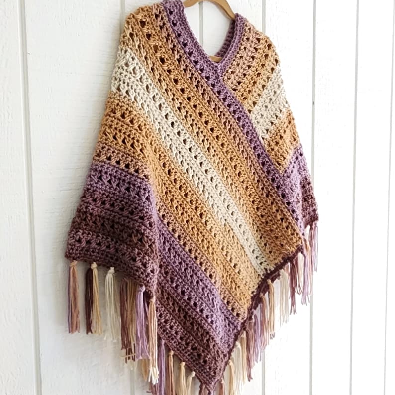 Puede incluir: Un poncho de ganchillo con cuello en V y un patr&oacute;n de rayas diagonales en tonos morado, beige y crema. La prenda est&aacute; confeccionada con un dise&ntilde;o de punto calado y terminada con largos flecos anudados a lo largo del dobladillo inferior.