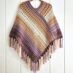 Puede incluir: Un poncho de ganchillo con cuello en V y un patr&oacute;n de rayas diagonales en tonos crema, bronceado, marr&oacute;n y morado. La prenda est&aacute; terminada con largos flecos de hilo multicolor a lo largo de los bordes inferiores, creando un dobladillo en punta.