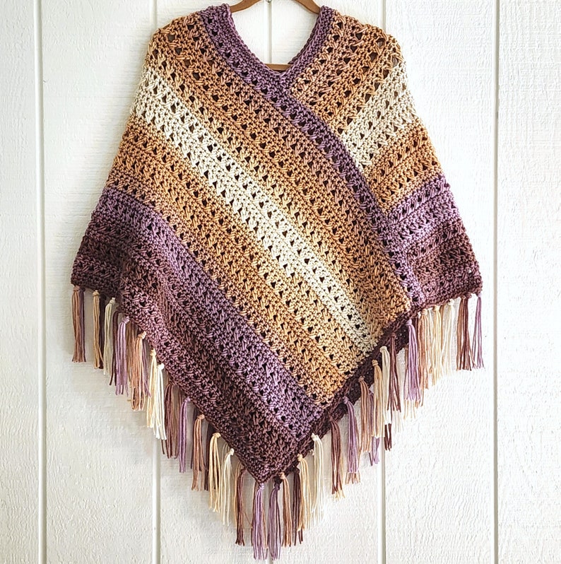 Puede incluir: Un poncho de ganchillo con cuello en V y un patr&oacute;n de rayas diagonales en tonos crema, bronceado, marr&oacute;n y morado. La prenda est&aacute; terminada con largos flecos de hilo multicolor a lo largo de los bordes inferiores, creando un dobladillo en punta.