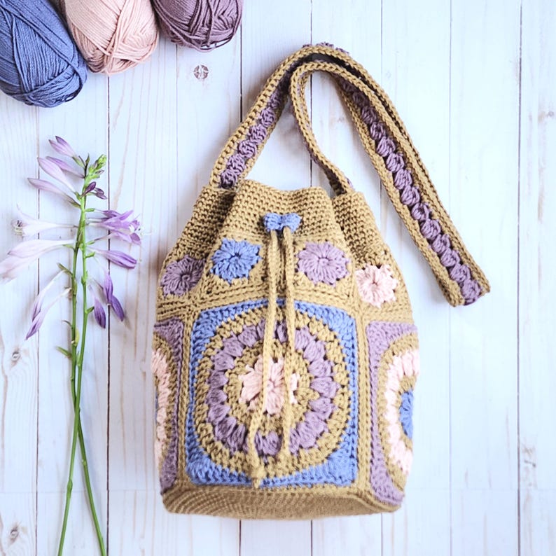 K&ouml;nnte beinhalten: Handgefertigte H&auml;keltasche in Beige, Blau und Rosa. Die Tasche hat ein Blumenmuster auf der Vorder- und den Seiten, einen Kordelzugverschluss und einen langen Riemen. Die Tasche ist aus Garn gefertigt und ein einzigartiges, stilvolles Accessoire.