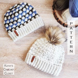 May include: Two handmade crochet beanies. One beanie is white with a faux fur pom-pom, and the other is blue, white, and brown. Both have a ribbed brim and a small leather tag. The text "Kathy's Crochet Closet" is in the lower left corner.