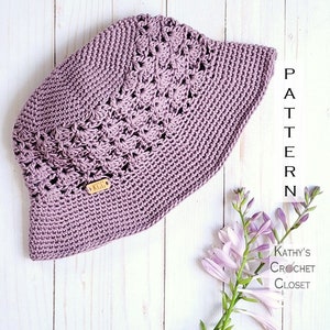 May include: A crocheted sun hat in a light purple color with a delicate openwork pattern. The hat is lying on a white wooden surface with a small wooden tag that says "KCG".