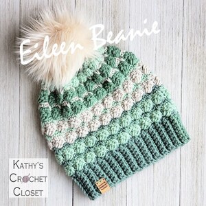 May include: A green, white and cream crocheted beanie with a fluffy pom pom on top. The beanie has a label that says "Kathy's Crochet Closet".