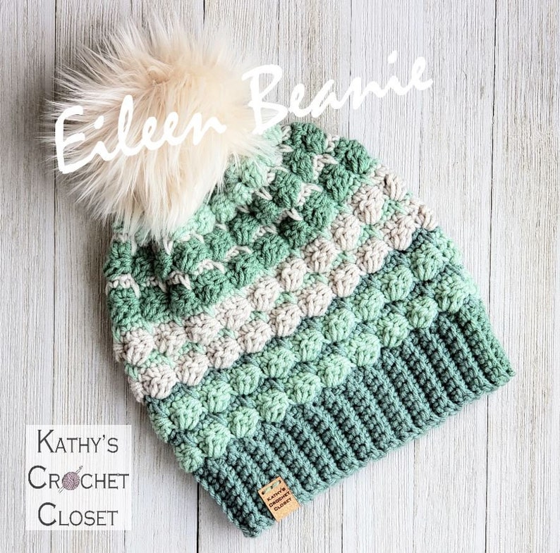 May include: A green, white and cream crocheted beanie with a fluffy pom pom on top. The beanie has a label that says "Kathy's Crochet Closet".