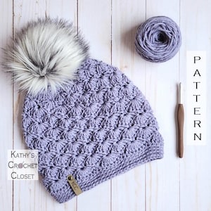 Puede incluir: Un gorro de crochet morado con un pompón blanco. El gorro está hecho con un punto texturizado y tiene una pequeña etiqueta de cuero marrón con las letras "KCC". El gorro está sobre una superficie de madera blanca con un ovillo de lana morado y un ganchillo.