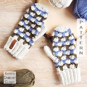 May include: A pair of handmade crochet mittens with a textured pattern in blue, white, and olive green. The cuffs and tops of the mittens are light gray. A small leather tag with the initials "KCC" is attached to one mitten. Yarn skeins are in the background.