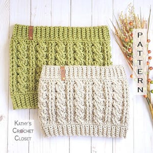 Puede incluir: Dos calentadores de cuello de ganchillo, uno de color verde claro y otro de color beige. Ambos tienen un patrón de punto de cable y una etiqueta de cuero con las letras "KCC" en ellos. El texto "PATTERN" es visible en el fondo.