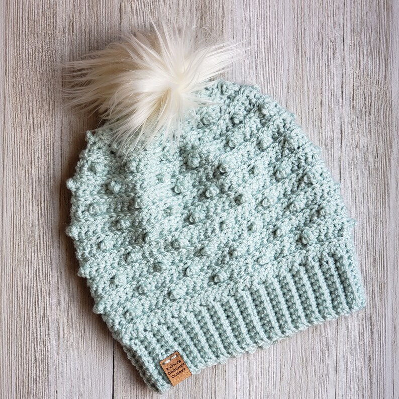 May include: A light green crocheted beanie with a white pom pom on top. The beanie has a textured stitch pattern and a brown leather tag with the text "Kait's Crocheted Creations" on it.