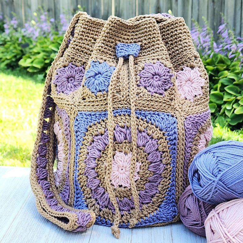 K&ouml;nnte beinhalten: Handgefertigter H&auml;kel-Rucksack mit Kordelzug in Beige-, Lila-, Blau- und Rosat&ouml;nen. Die Tasche hat ein Blumenmuster und einen Kordelzugverschluss. Der Riemen ist passend geh&auml;kelt. Garnkn&auml;uel in passenden Farben befinden sich im Vordergrund.