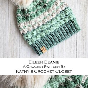 May include: A green and white crochet beanie with a fluffy pom pom. The beanie has a textured stitch pattern and is labelled "Kathy's Crochet Closet".
