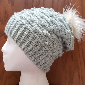 May include: A light blue crocheted beanie with a white pom pom. The beanie has a textured stitch pattern.