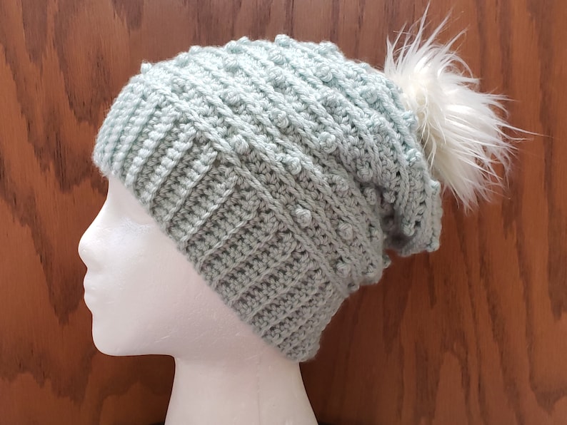May include: A light blue crocheted beanie with a white pom pom. The beanie has a textured stitch pattern.