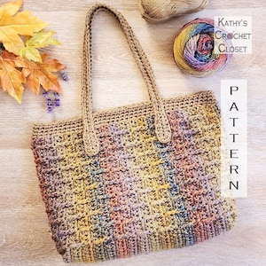 May include: A handmade crochet tote bag with a textured stitch pattern. The bag features a neutral beige color with colorful accents of pink, yellow, and blue. The bag has two handles and the text "Kathy's Crochet Closet" and "Pattern" are visible.