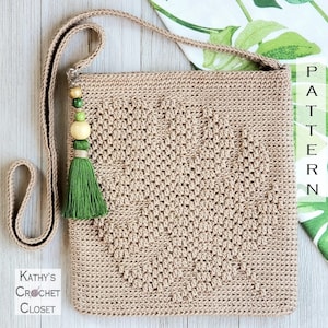 Puede incluir: Un bolso de hombro de color beige tejido a crochet con un borla verde y cuentas de madera. El bolso tiene un diseño en forma de corazón y el texto "Kathy's Crochet Closet" es visible en la esquina inferior izquierda.
