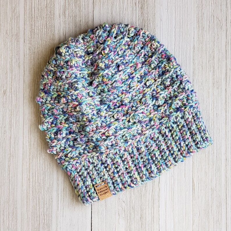 May include: A colorful crocheted beanie hat with a textured pattern. The hat has a brown leather tag with the text "Kathy's Crochet Closet".