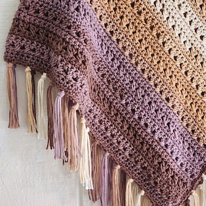 Puede incluir: Un chal triangular de ganchillo con un patr&oacute;n de color degradado que transita de ciruela intenso a marr&oacute;n c&aacute;lido y crema. El tejido tiene un dise&ntilde;o de punto calado y est&aacute; rematado con largas borlas de hilo multicolor en el borde.