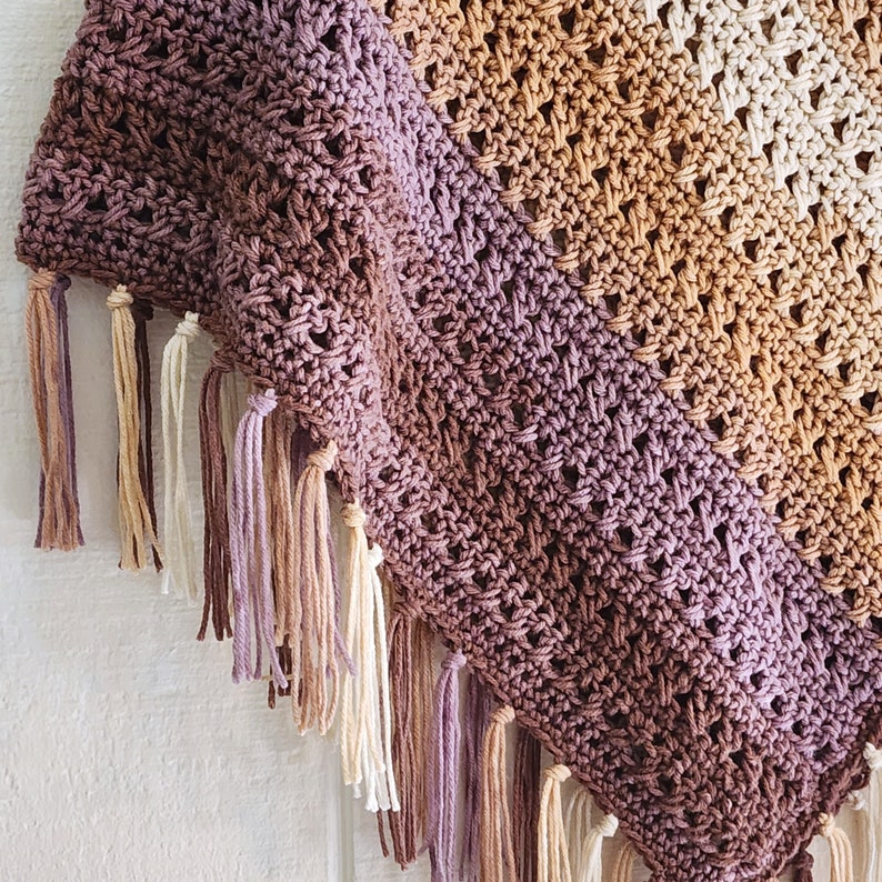 Puede incluir: Un chal triangular de ganchillo con un patr&oacute;n de color degradado que transita de ciruela intenso a marr&oacute;n c&aacute;lido y crema. El tejido tiene un dise&ntilde;o de punto calado y est&aacute; rematado con largas borlas de hilo multicolor en el borde.
