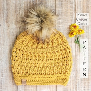 May include: A mustard yellow crochet beanie with a faux fur pom pom. The beanie has a textured stitch pattern and a small brown leather tag with the text "Kathy's Crochet Closet" on it.