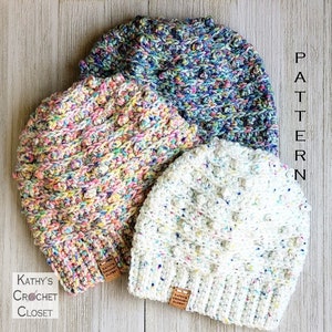 Op de afbeelding: Drie gebreide mutsen in verschillende kleuren: wit, roze en blauw met witte stippen. De mutsen hebben een textuur, een hobbelig ontwerp. De mutsen liggen op een houten oppervlak. De tekst "PATTERN" is zichtbaar aan de rechterkant van de afbeelding.