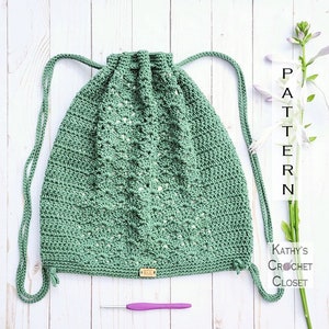 以下が含まれることがあります： 星形のステッチパターンが施された緑色のクロシェの巾着袋。袋には「KCD」という文字が書かれた木製のタグが付いています。袋は白い木製の表面に置かれ、紫色の編み針が置かれています。