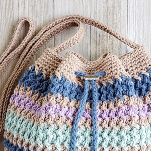 Pu&ograve; includere: Una borsa a cordoncino all'uncinetto con un motivo a righe marroni, blu, viola e verdi. La borsa ha una tracolla marrone e una chiusura in metallo argentato.