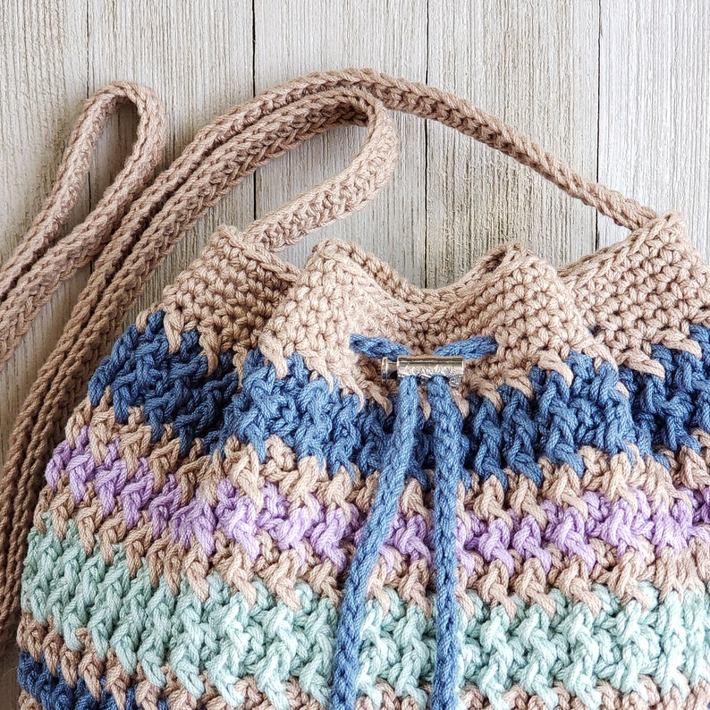 Pu&ograve; includere: Una borsa a cordoncino all'uncinetto con un motivo a righe marroni, blu, viola e verdi. La borsa ha una tracolla marrone e una chiusura in metallo argentato.