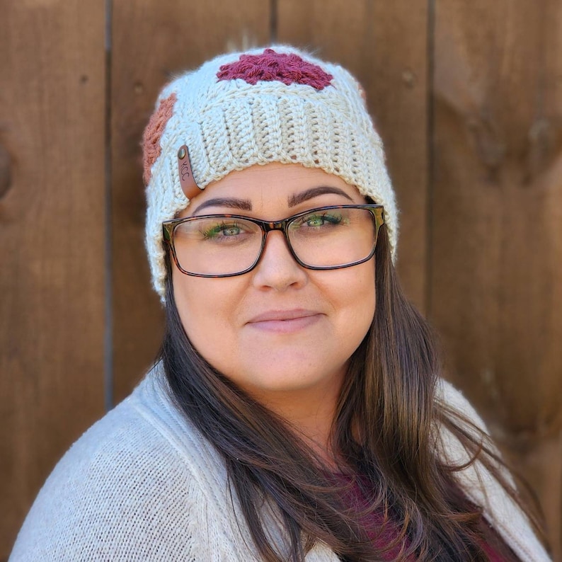 May include: A woman wearing a white crocheted beanie with brown and red flower accents. The beanie has a brown leather tag with the word "Cozy" on it. She is also wearing glasses.