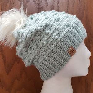 May include: A light blue crocheted beanie with a white pom pom on top. The beanie has a textured stitch pattern and a brown leather tag that says "Katie's Crochet Closet".