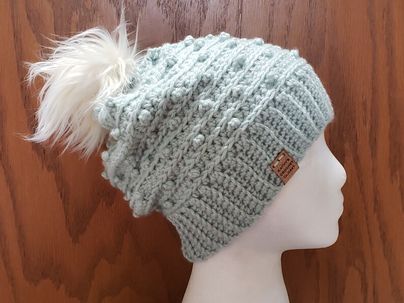 May include: A light blue crocheted beanie with a white pom pom on top. The beanie has a textured stitch pattern and a brown leather tag that says "Katie's Crochet Closet".