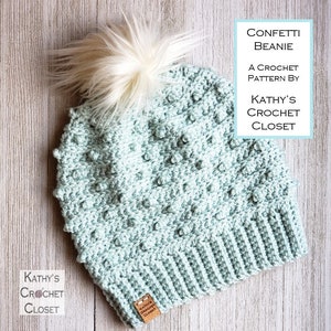May include: A light blue crochet beanie with a white pom pom. The beanie has a textured stitch pattern and a fold-over brim. The beanie is labeled "Confetti Beanie" and "A Crochet Pattern by Kathy's Crochet Closet".