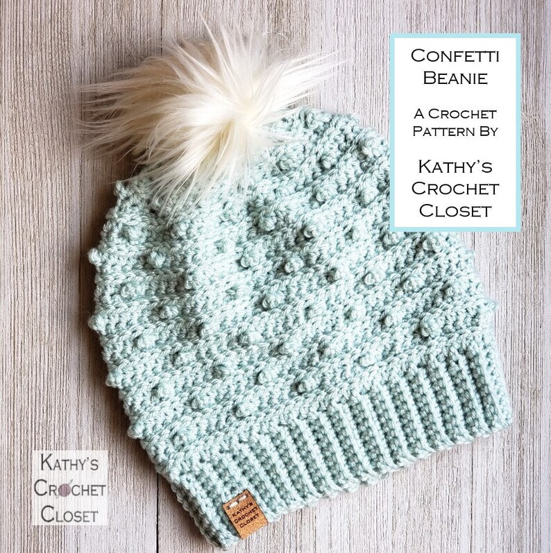 May include: A light blue crochet beanie with a white pom pom. The beanie has a textured stitch pattern and a fold-over brim. The beanie is labeled "Confetti Beanie" and "A Crochet Pattern by Kathy's Crochet Closet".