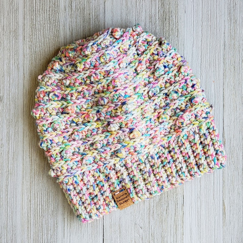 May include: A colorful crocheted hat with a multi-colored yarn. The hat has a textured design and a small brown tag that reads "Kathy's Crochet Closet".