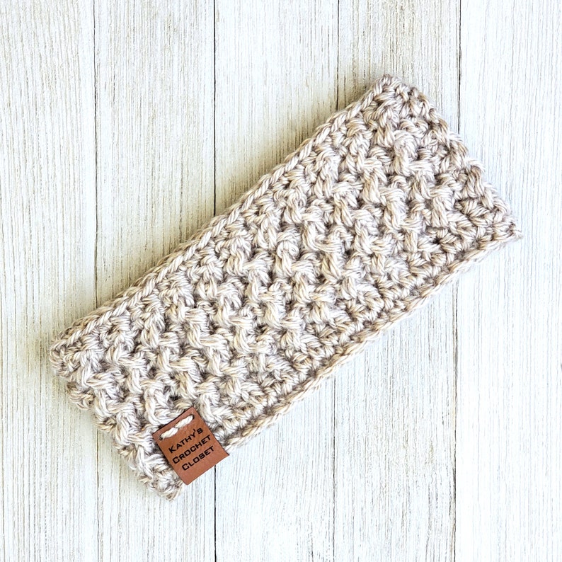 Op de afbeelding: Een beige gehaakt hoofdband met een bruin leren label waarop "Kathy's Crochet Closet" staat.