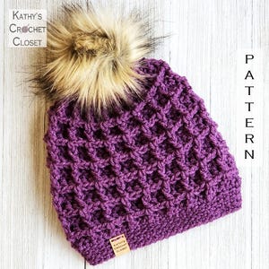 May include: A purple crocheted beanie with a waffle stitch texture and a large, fluffy beige faux fur pom-pom on top. A small rectangular wooden tag labeled Kathy's Crochet Closet is attached to the bottom edge. The text PATTERN is visible.