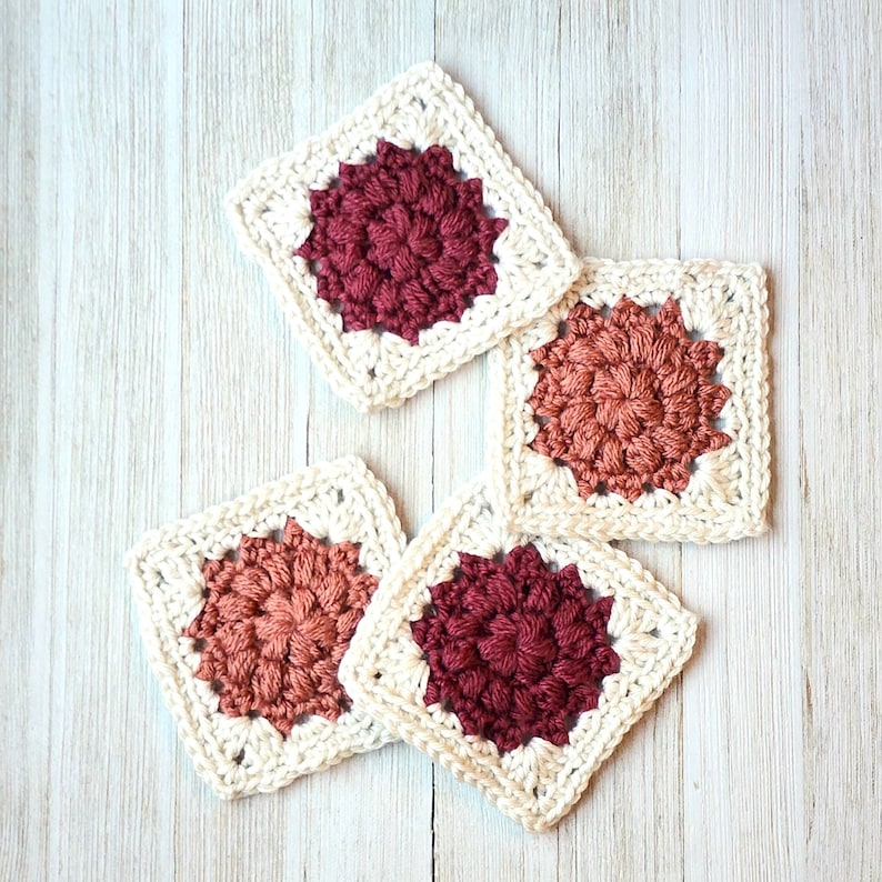 May include: Four crocheted squares with a white border and a red, pink, or orange center. The squares are made with a textured stitch.