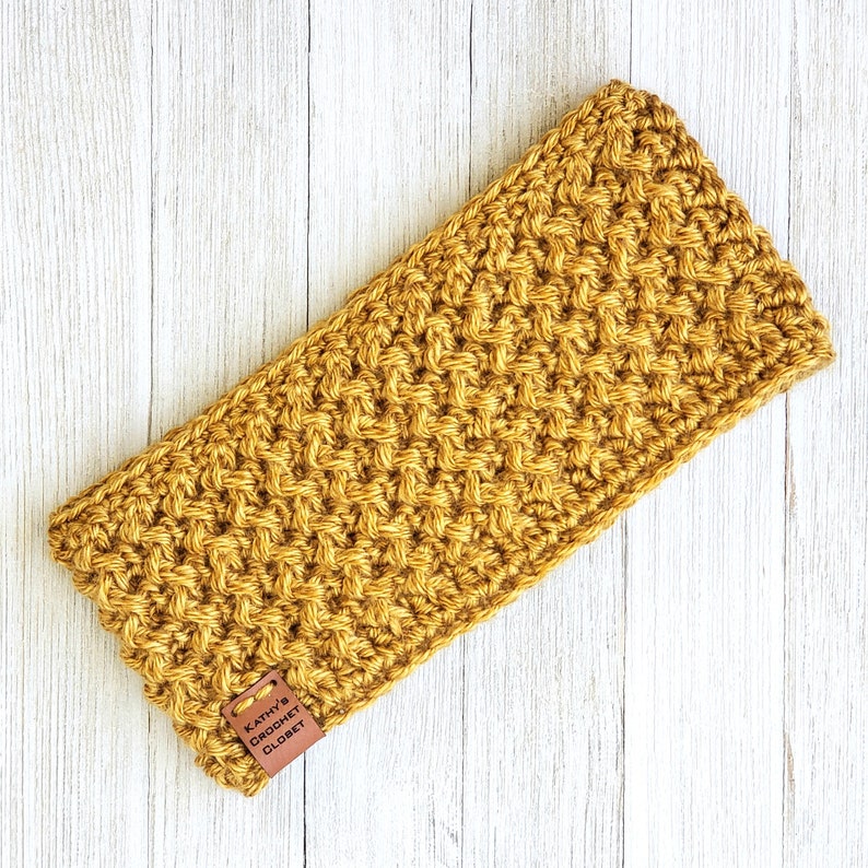 Op de afbeelding: Een mosterdgele gehaakte hoofdband met een bruin leren label waarop "Kathy's Crochet Closet" staat.