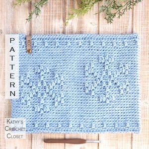 May include: A light blue crochet cowl with a textured pattern of hearts. The cowl is on a wooden surface with green foliage.