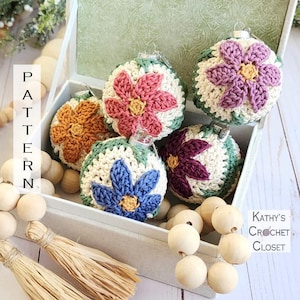 May include: Five crocheted ornaments in a box with wooden beads. The ornaments are in the shape of a flower with different colored petals. The colors are purple, pink, blue, orange, and green.