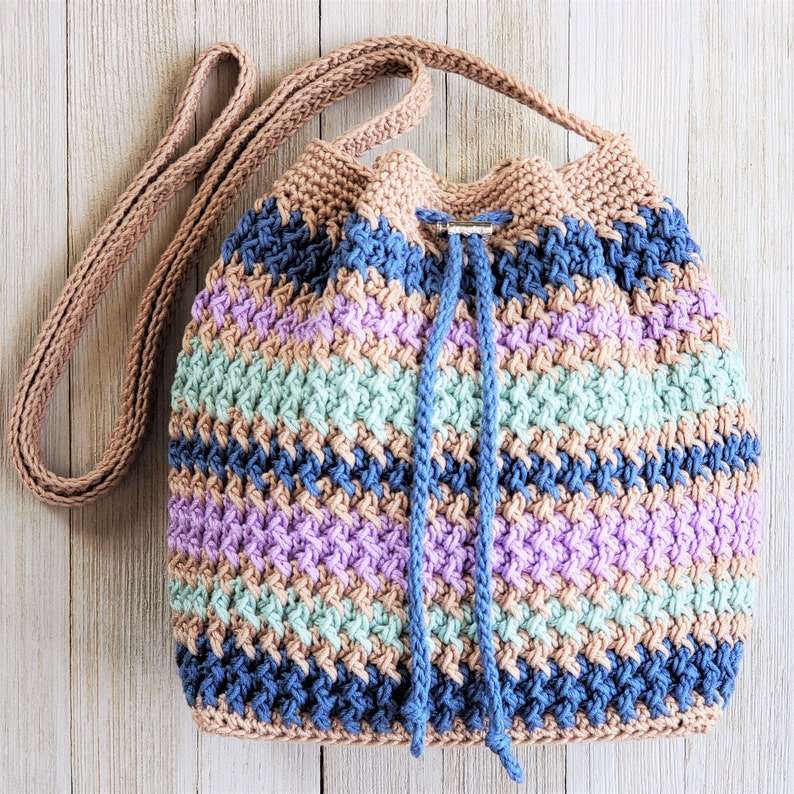 Pu&ograve; includere: Una borsa a cordoncino all'uncinetto con una lunga tracolla. La borsa &egrave; realizzata con una variet&agrave; di colori, tra cui blu, viola, verde e marrone. La borsa &egrave; perfetta per trasportare piccoli oggetti.