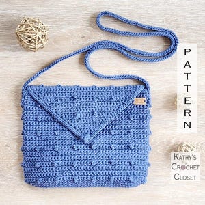 Könnte beinhalten: Handgefertigte blaue Häkeltasche mit Klappe und strukturiertem Noppenmuster. Die Tasche hat einen langen Riemen und ein kleines Holzschild. Der Text "PATTERN" und "Kathy's Crochet Closet" sind sichtbar.