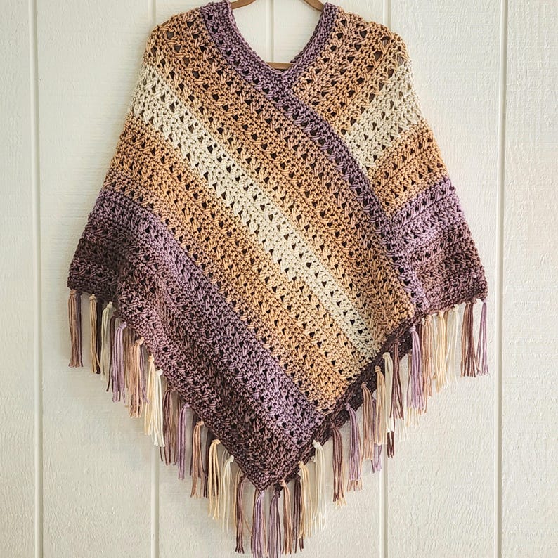 Puede incluir: Un poncho de ganchillo con cuello en V que presenta un patr&oacute;n de rayas diagonales en tonos morado, tostado, crema y marr&oacute;n. La prenda est&aacute; confeccionada con un dise&ntilde;o de punto abierto y rematada con largos flecos multicolores en el borde.