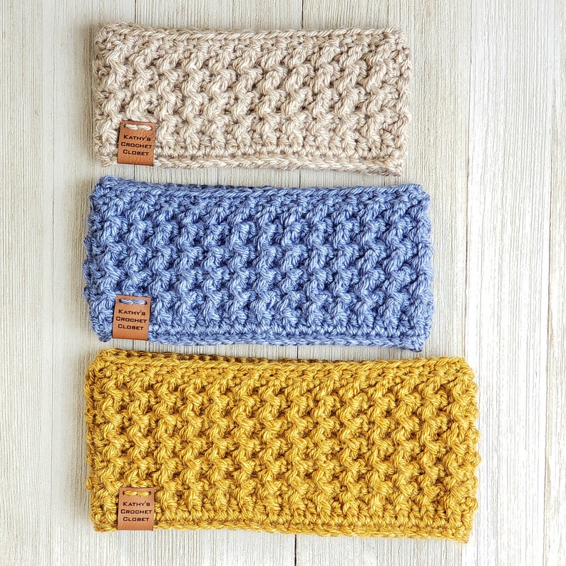 Op de afbeelding: Drie gehaakte hoofdbanden in beige, blauw en mosterdgeel. Elke hoofdband heeft een bruin lederen label met de tekst "Kathy's Crochet Closet".