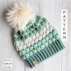 May include: A green, white and grey crocheted beanie with a fluffy white pom pom on top. The beanie has a textured stitch pattern and a brown leather tag with the text "Kathy's Crochet Closet".