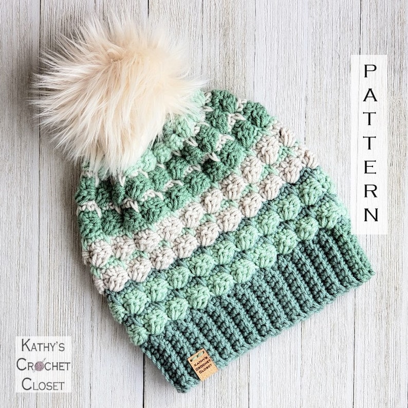 May include: A green, white and grey crocheted beanie with a fluffy white pom pom on top. The beanie has a textured stitch pattern and a brown leather tag with the text "Kathy's Crochet Closet".