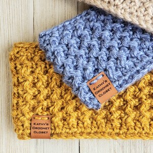 Op de afbeelding: Twee gebreide hoofdbanden, een mosterdgeel en een lichtblauw, met bruine leren labels waarop "Kathy's Crochet Closet" staat.