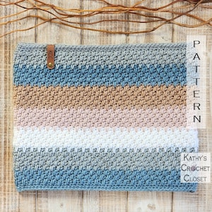 May include: A crocheted cowl with a striped pattern in shades of blue, gray, beige, pink, and white. The cowl has a leather tag with the initials "KCC" on it. The cowl is lying on a wooden surface.