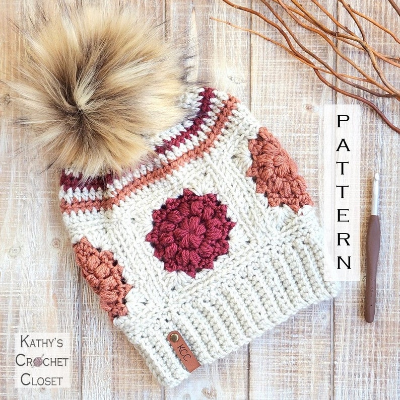 May include: A crocheted beanie hat with a faux fur pom pom. The hat is made in a cream color with a red, orange, and brown geometric pattern. The hat has a leather tag with the initials "KCC".