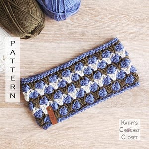 May include: A handmade blue, white, and olive green crochet headband with a textured pattern. The headband has a small leather tag with the initials "KCC". The word "PATTERN" is printed vertically on the left side of the image. The words "Kathy's Crochet Closet" are printed on the right side.